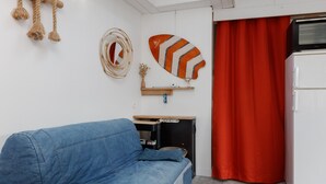 Un espacio habitable pequeño con un sofá azul, una mesita de madera, un refrigerador blanco y una cortina roja.