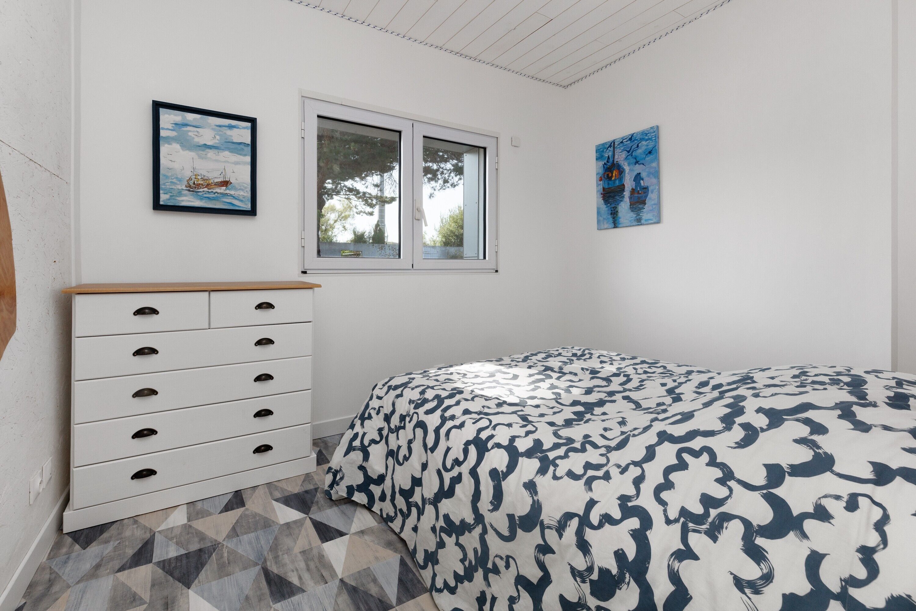 Un dormitorio con cama, cómoda, ventana y dos cuadros en la pared.