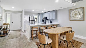 A modern kitchen with white cabinets, a central island, and a dining area with a wooden table and wicker chairs. - Sunset Rentals | Queens Grant 616 | Dog Friendly (Hilton Head Island)