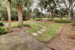 A landscaped garden with a stone pathway, palm trees, and a golf course in the background. - Sunset Rentals | Queens Grant 616 | Dog Friendly (Hilton Head Island)