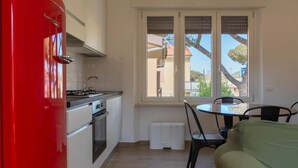 Uma cozinha compacta com um eletrodoméstico vermelho, um espaço de refeições com mesa e cadeiras, e uma janela com vista para edifícios e árvores.