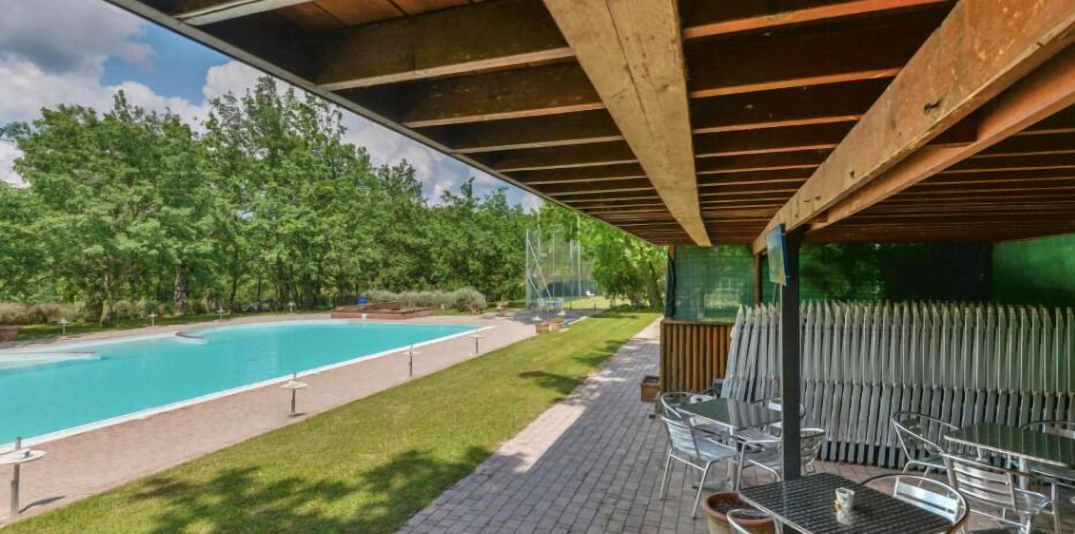 A covered outdoor area with a swimming pool, chairs, and a view of a forest.