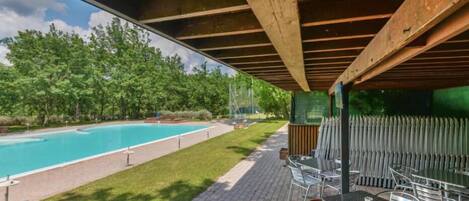 A covered outdoor area with a swimming pool, chairs, and a view of a forest.