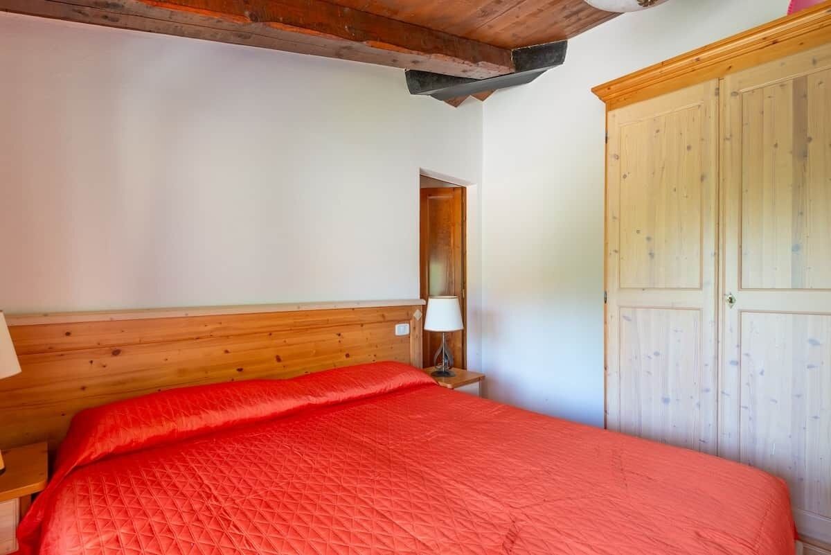 A bedroom with a wooden headboard, a red quilted bedspread, a bedside lamp, and a wooden wardrobe.