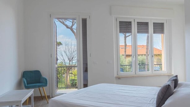 Een slaapkamer met een bed, een stoel, een tafel en een raam met uitzicht op gebouwen en bomen.