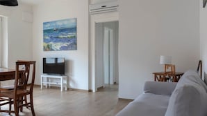 Een woonkamer met een televisie, een schilderij, een bank en een eethoek met stoelen en een tafel.