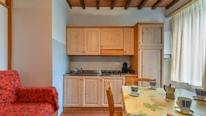 A compact kitchen with wooden cabinets, a dining table with a floral tablecloth, and a red sofa. - To complete (Castel del Piano)