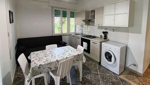 A compact kitchen with a dining area, a washing machine, and a window with shutters. - 454 Orchidee Mare (3-seater) (Marina di Grosseto)