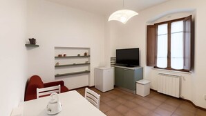 A compact living space with a dining table, chairs, a TV, a small fridge, and a window with blinds. - Casa Maria Amiata: A Cozy House Amid Nature and Comfort (San Lorenzo)