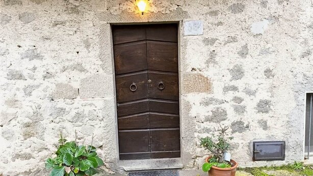 A rustic stone building with a dark wooden door, two potted plants, and a small light fixture above the door. - Casa Maria Amiata: A Cozy House Amid Nature and Comfort (San Lorenzo)