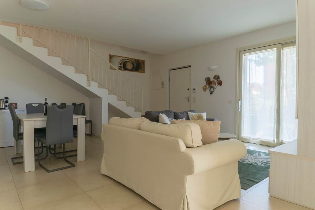 A living room with a sofa, a dining table with chairs, a staircase, and a sliding glass door.