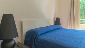 A bed with a blue quilt, a black lamp on a bedside table, and a window with curtains. - To complete (Grosseto)