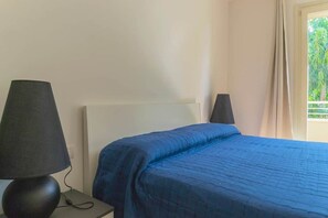 A bed with a blue quilt, a black lamp on a bedside table, and a window with curtains. - To complete (Grosseto)
