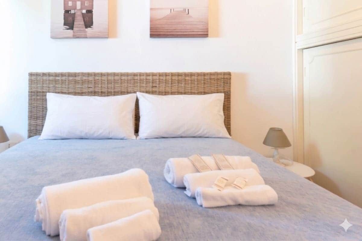 Een opgemaakt bed met witte kussens en handdoeken, een rieten hoofdbord en twee aan de muur hangende foto's.