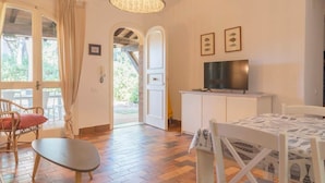Ein Zimmer mit Fernseher, Esstisch, Lampenschirm aus Weidengeflecht und Blick durch eine offene Tür.