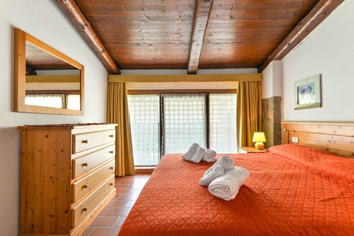 Una camera da letto con una cassettiera in legno, un letto con copripiumino arancione, uno specchio, una lampada e una finestra con tende.