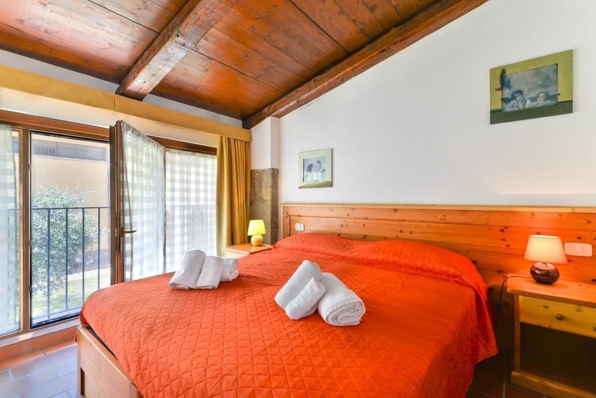 Habitación de hotel con techo de madera, una cama grande con colcha naranja, dos toallas enrolladas, una mesita de noche con lámpara y una ventana con cortinas.