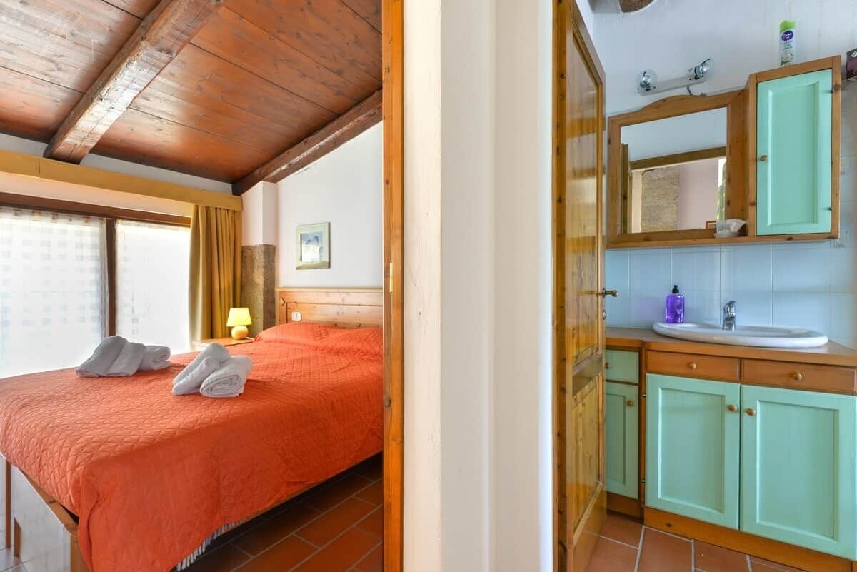 Une chambre avec un plafond en bois, un lit recouvert d’une courtepointe orange, une table de chevet avec une lampe, et une salle de bain avec un lavabo et des placards.