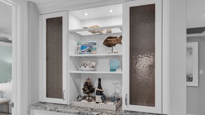 A white cabinet with shelves containing decorative items, a small Christmas tree, and a bottle of wine. - South End Waterfront Condo | Walk to Beach (Marco Island)