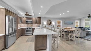 A modern kitchen with built-in appliances, a dining area with a large island, and a living room with a sofa and a painting. - South End Waterfront Condo | Walk to Beach (Marco Island)