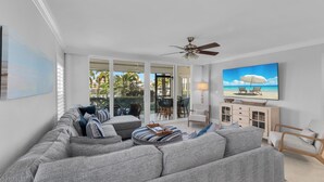 A modern living room with a large grey sectional sofa, a ceiling fan, and a flat-screen TV mounted on the wall. - South End Waterfront Condo | Walk to Beach (Marco Island)