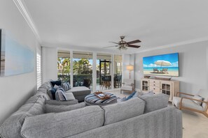 A modern living room with a large grey sectional sofa, a ceiling fan, and a flat-screen TV mounted on the wall. - South End Waterfront Condo | Walk to Beach (Marco Island)