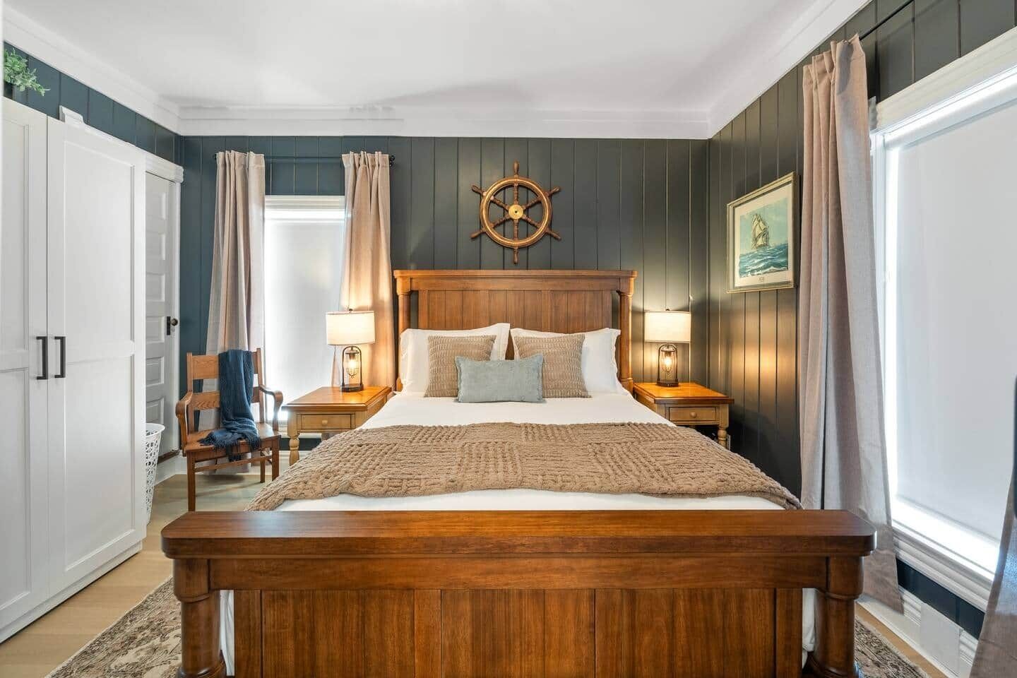 A bedroom with a wooden bed, two bedside tables with lamps, a ship's wheel decoration, and a framed picture on the wall.