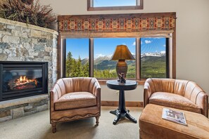 A room with a stone fireplace, leather armchairs, a round table with a lamp, and a large window with a mountain view.