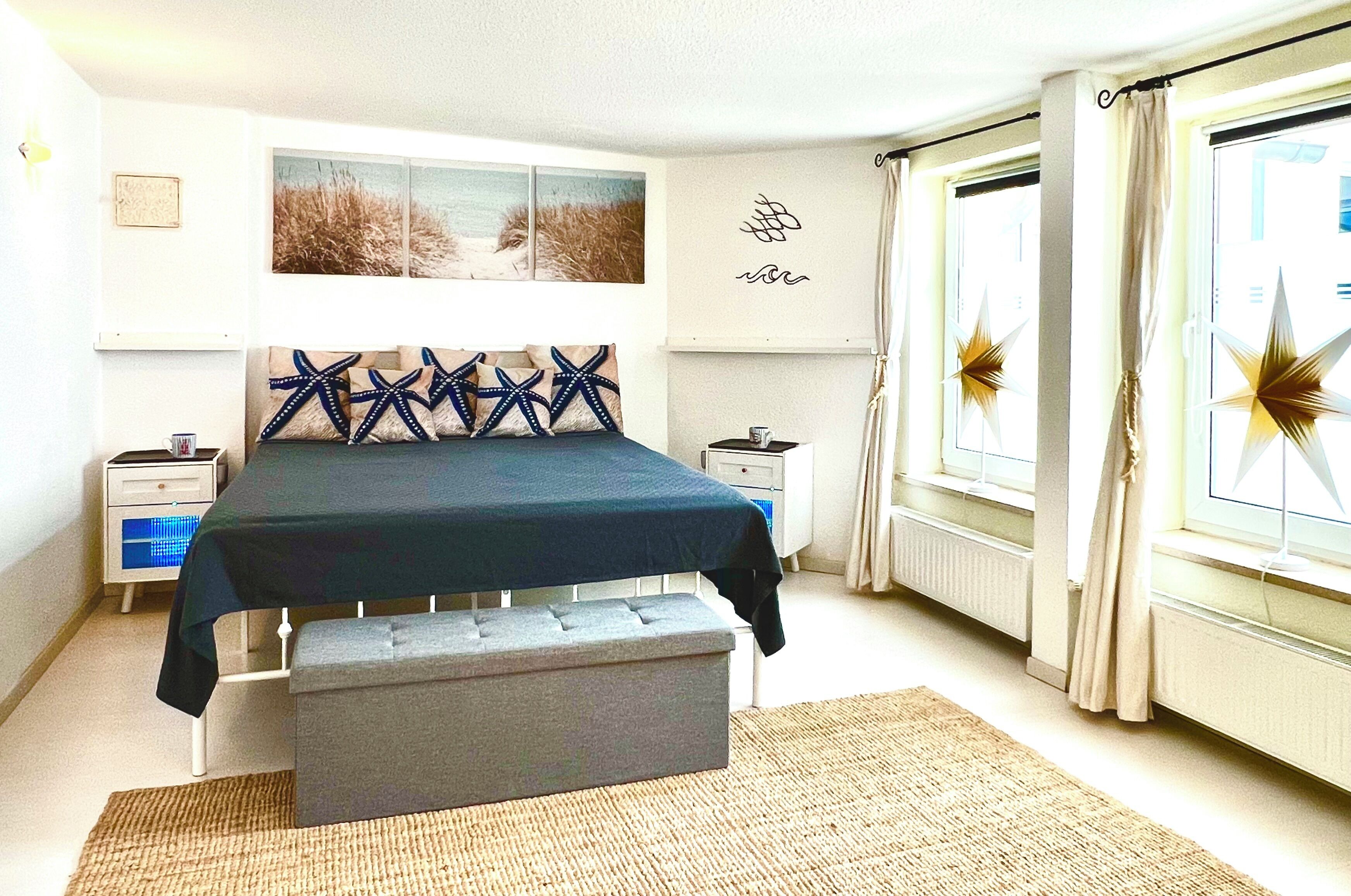 A bedroom with a bed, bedside tables, a bench, a window with curtains, and wall art.