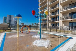 A water play area with multiple water jets and colorful rings, surrounded by a fenced pool area and a multi-story building with balconies. - Phoenix X 1506 3 Bedroom Condo (Orange Beach)