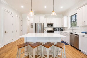 A modern kitchen with a central island, bar stools, stainless steel appliances, and pendant lighting. - Endless Summer 161 4 Bedroom Home (Gulf Shores)