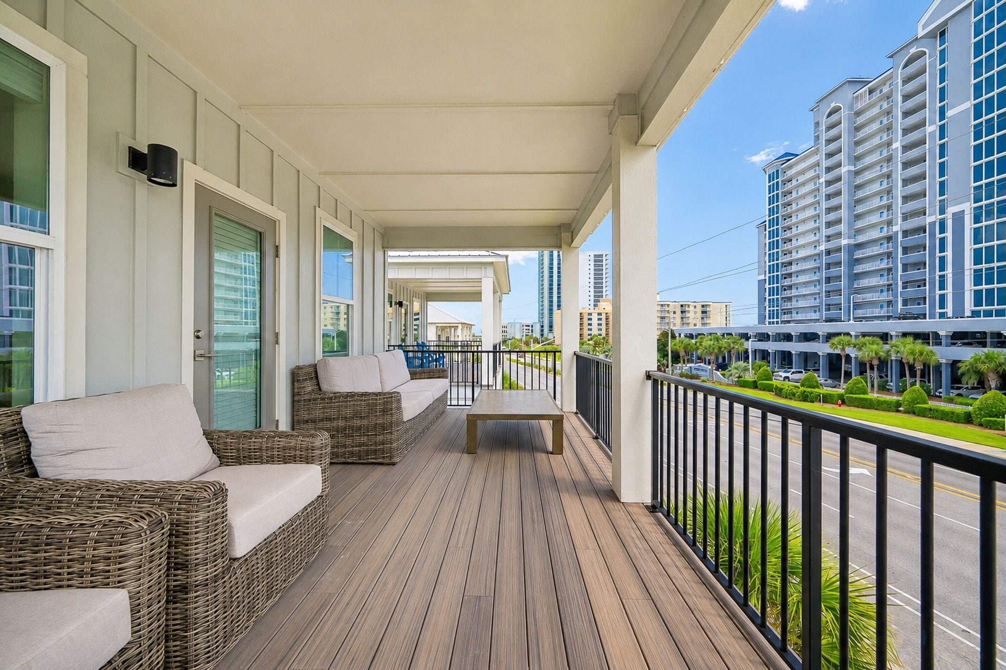 A covered outdoor balcony with wicker furniture and a bench, overlooking a street and buildings.