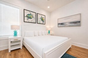 A bedroom with a large bed, two bedside tables, a window with blinds, and artwork on the walls. - Endless Summer 161 4 Bedroom Home (Gulf Shores)
