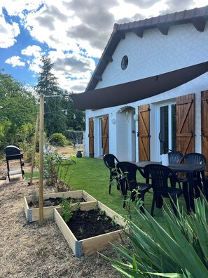 Una casa con fachada blanca, contraventanas de madera, un jardín con jardineras elevadas y un patio con sillas y una mesa.