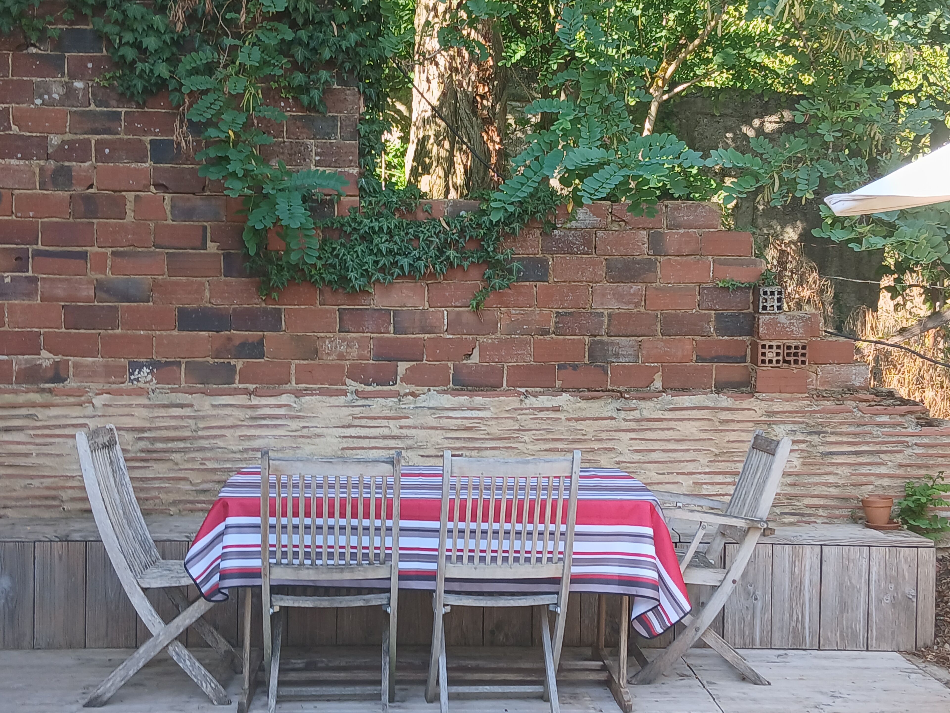 A brick wall with climbing plants, a wooden table with striped tablecloth, and two wooden chairs.