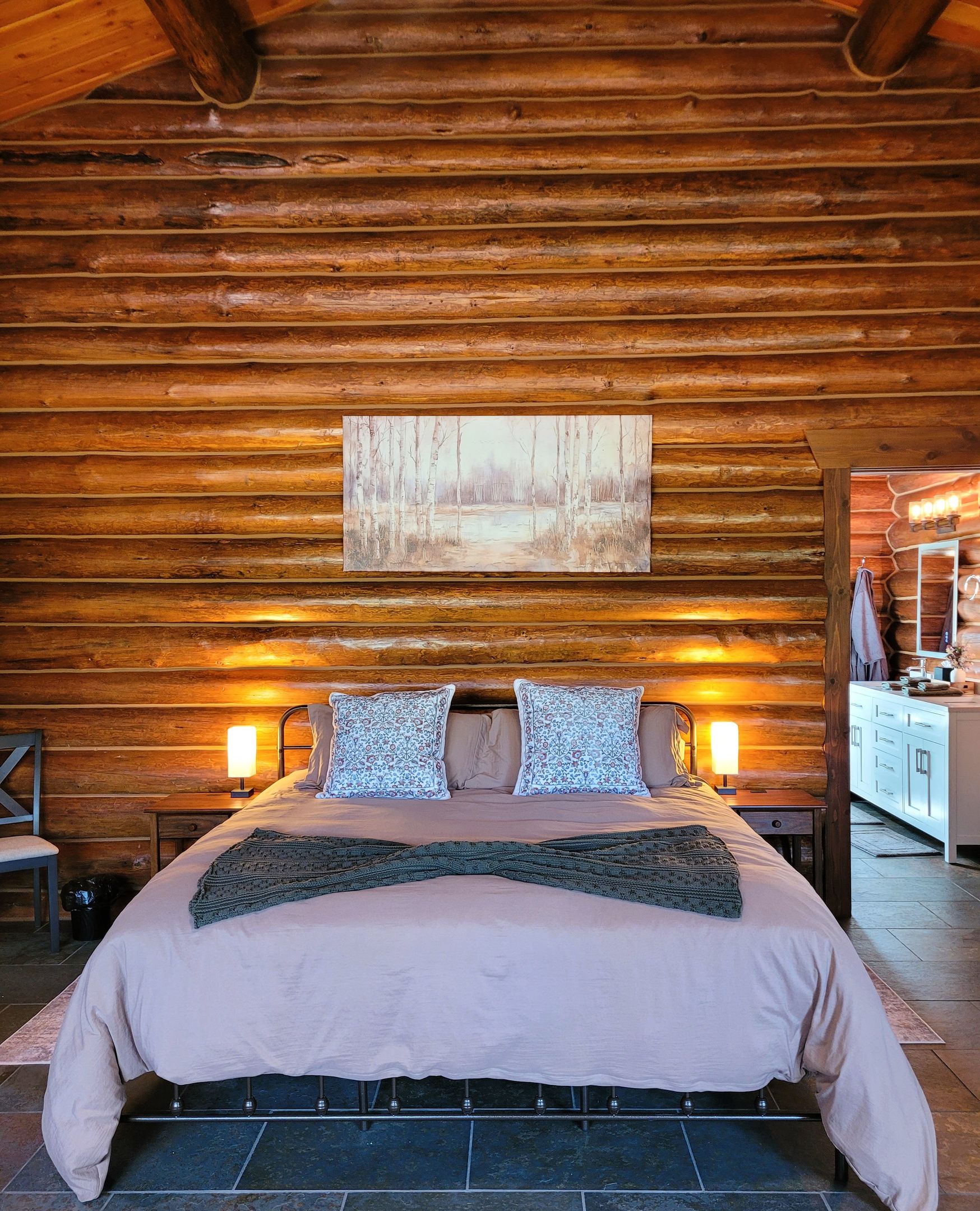 A log cabin bedroom with a bed, pillows, a painting, and a bathroom visible.