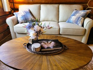 Una mesa de centro redonda de madera con un jarrón decorativo, una vela y un libro titulado 'The Hidden Life of Wolves' frente a un sofá color beige con almohadones estampados.
