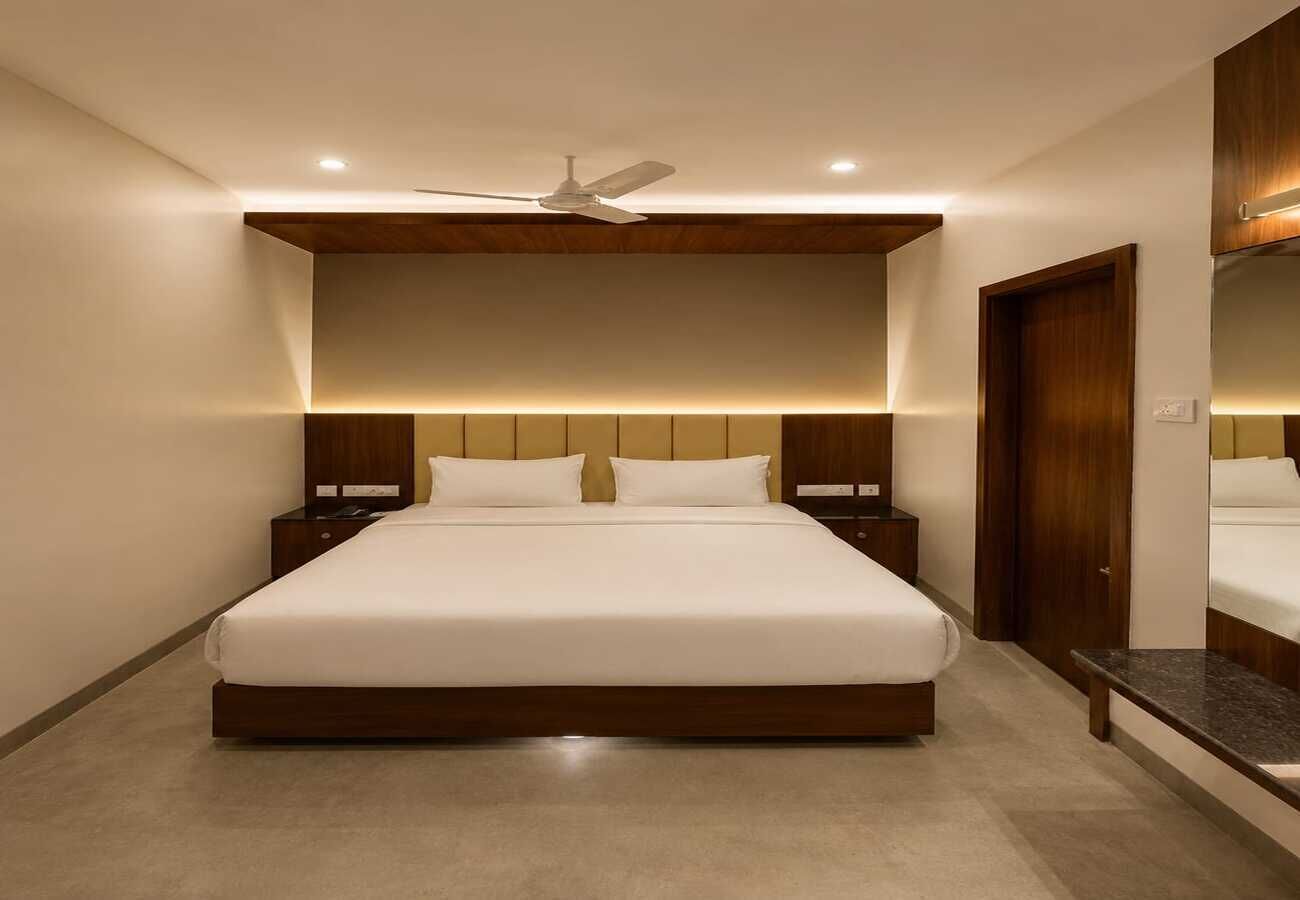 A modern hotel bedroom with a large bed, wooden headboard, bedside tables, a ceiling fan, and a large mirror.