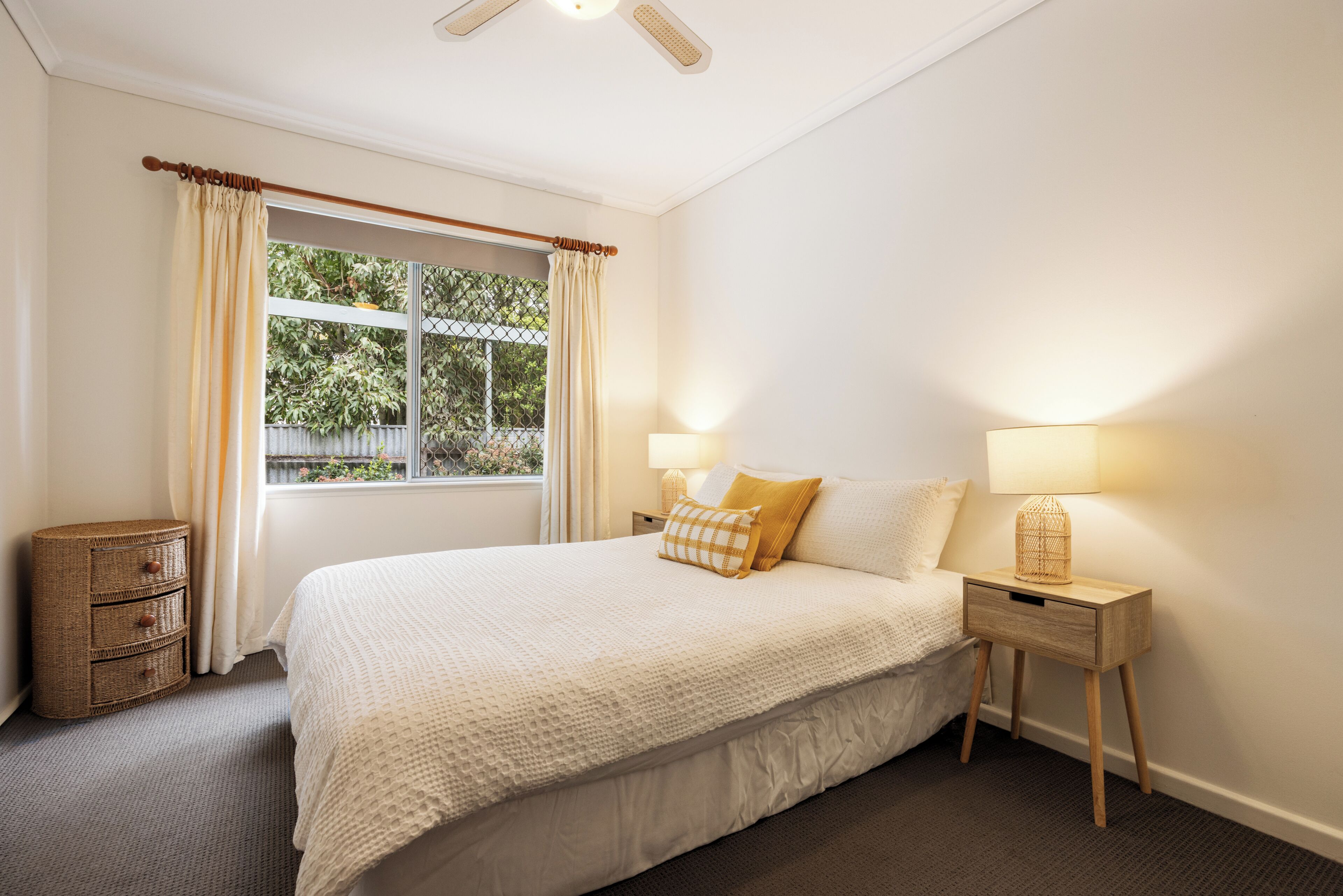 A bedroom with a bed, two bedside tables, a nightstand, a window with curtains, and a view of greenery outside.