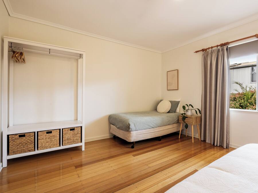 Quarto com cama, mesa de cabeceira com planta, janela com cortinas e guarda-roupa com roupas penduradas.