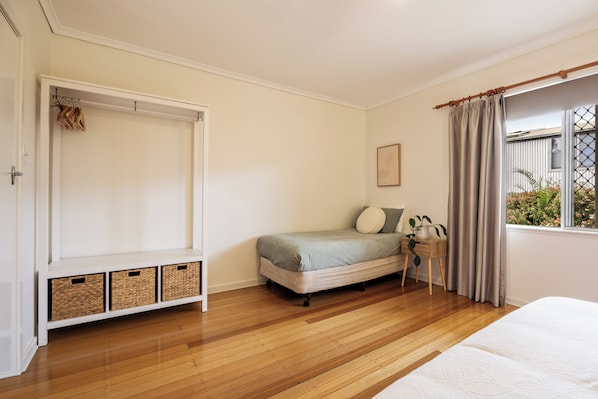 A bedroom with a bed, a bedside table with a plant, a window with curtains, and a wardrobe with hanging clothes.