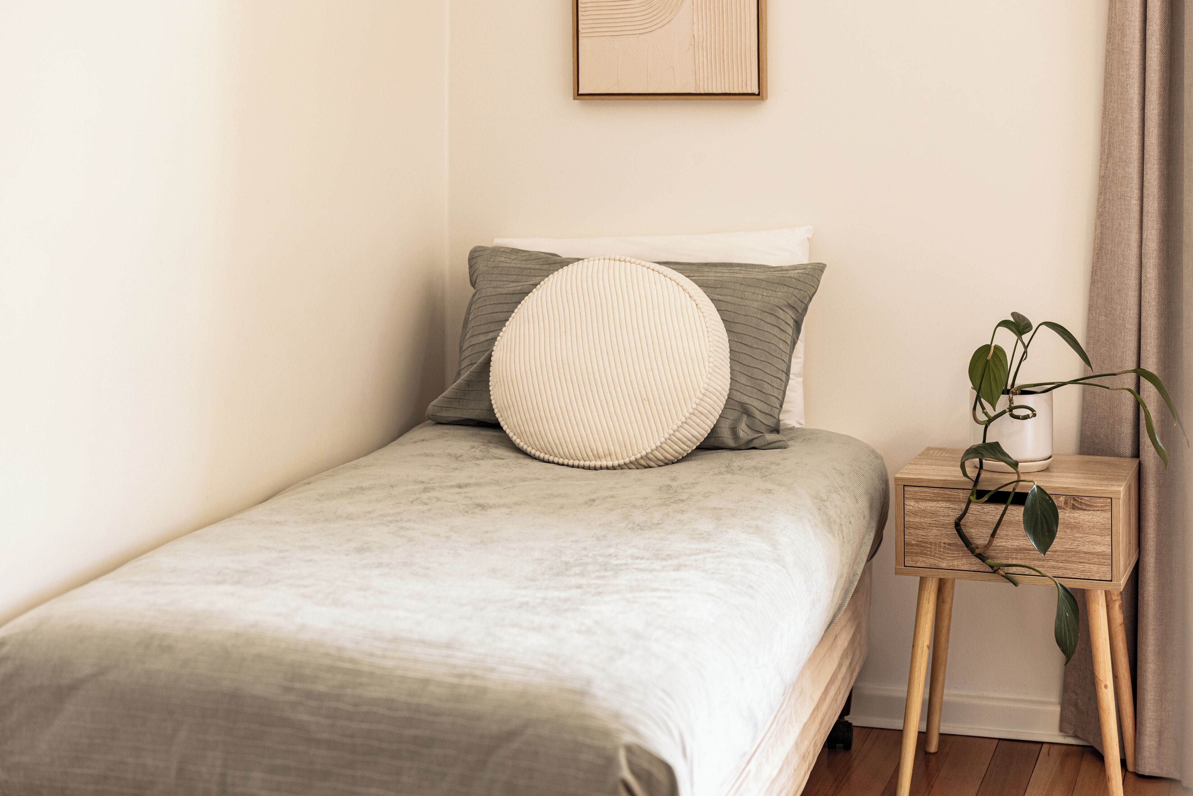 Una cama individual con una colcha gris, dos almohadas, un cojín redondo, una mesita de noche con una planta y un cuadro en la pared.
