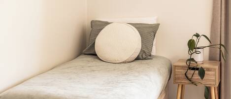 Una cama individual con una colcha gris, dos almohadas, un cojín redondo, una mesita de noche con una planta y un cuadro en la pared.