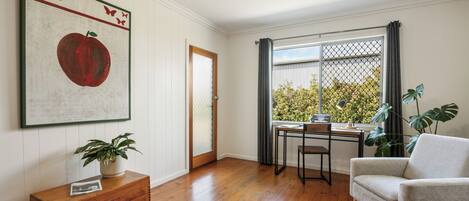 A room with a wooden floor, a desk with a chair, a window with a view of greenery, a plant on a wooden chest, and a framed picture on the wall.