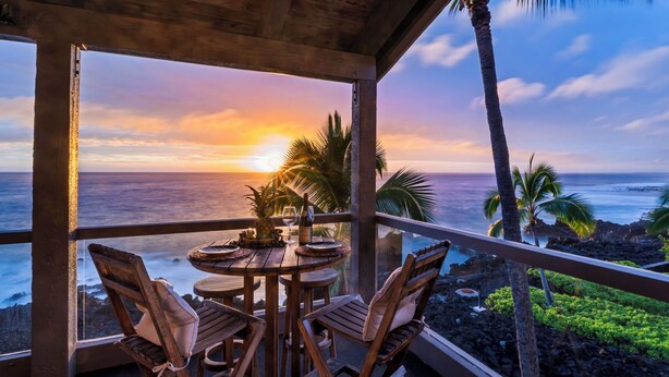 A wooden table with chairs and a bottle of wine, overlooking a sunset over the ocean with palm trees. - Kksr#1301 Direct Oceanfront, Best North Corner Top Floor Location (KAILUA KONA)