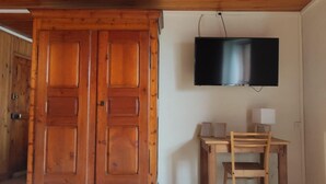 Una habitación con un armario de madera, un televisor montado en la pared, un escritorio de madera con una silla y un vaso de bebida en el suelo.