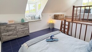 Ein Schlafzimmer mit schrägen Wänden, einer Holzkommode, einem Bett mit weißer Bettdecke und einem Fenster, das für natürliches Licht sorgt.
