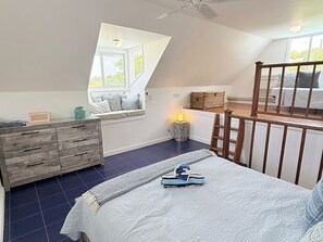 Ein Schlafzimmer mit schrägen Wänden, einer Holzkommode, einem Bett mit weißer Bettdecke und einem Fenster, das für natürliches Licht sorgt.