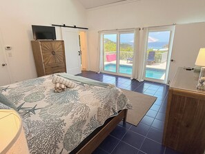 A bedroom with a bed, a television on a cabinet, a sliding glass door leading to a balcony with a view of the ocean. - Private 3 Bedroom Villa with Pool, Central A/C & Ocean Views in Chocolate Hole, St. John, USVI (St John)