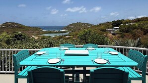Ein türkisfarben gedeckter Tisch mit Tellern, Besteck und Gläsern, mit Blick auf eine Küstenlandschaft mit Hügeln und einem Gewässer.